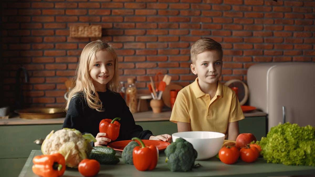Positive little girl and boy sitting at table with various vegetables and herbs while preparing vegetarian meal in loft kitchen with brick wall